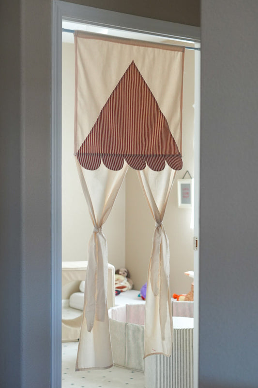 A kids playroom curtain with a striped red and beige triangle top and tie-backs, hanging in a doorway to a child's play area.