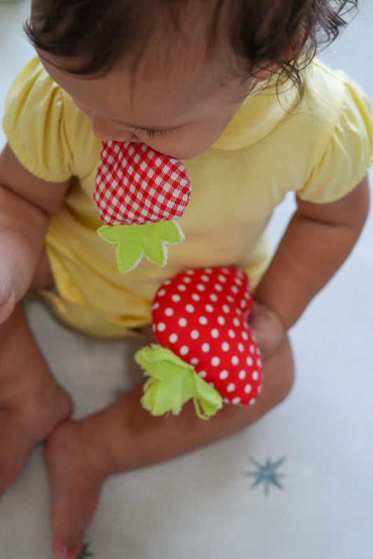 A baby wearing a yellow short-sleeve romper sits on a light cream play mat, holding a red strawberry-shaped fabric toy with white polka dots and green leaves, while touching another matching strawberry toy near their mouth.