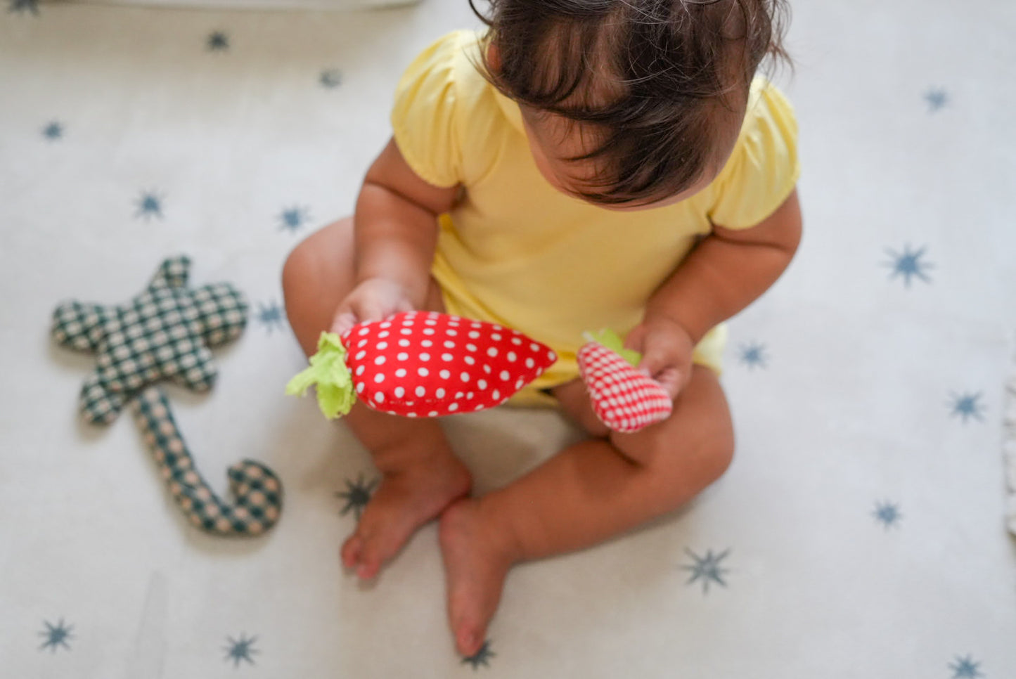 A baby in a yellow onesie sits on a white star-patterned floor mat, holding a red and white polka dot strawberry-shaped plush toy next to a green and white checkered plush toy.