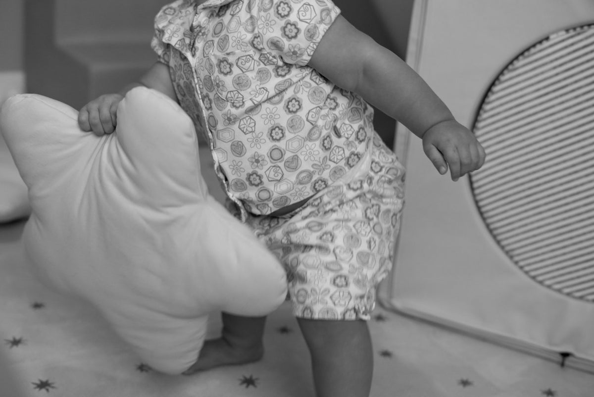 A baby wearing a short-sleeve patterned romper and matching shorts stands on a star-patterned floor mat, holding a large star shaped pillow next to a soft play toy.