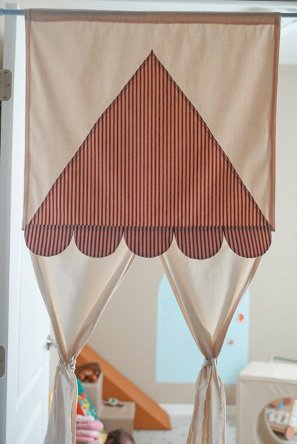 A fabric play curtain with a scalloped top edge and tie-on bottom, featuring a large triangle panel with vertical rust-red and cream stripes over a cream background.
