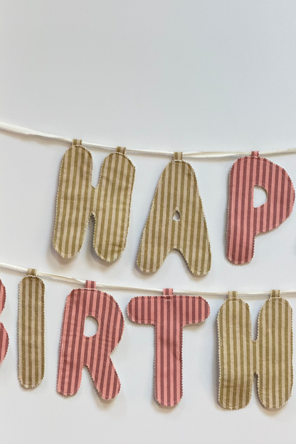 A close up of a fabric letter banner spelling 'HAPPY BIRTHDAY' hanging on a white string against a plain white background. The letters alternate between green and pink striped fabrics.