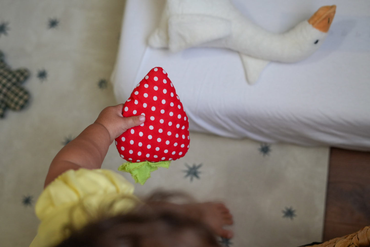A baby's hand holds a red strawberry-shaped plush toy with white polka dots and a green leafy stem. The baby is wearing a yellow top, and a white goose plush toy is visible on a white bed in the background.