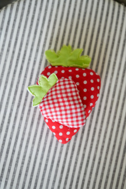 A small strawberry-shaped fabric cushion or pillow with a red and white gingham heart on top, set on a gray and white striped fabric background.