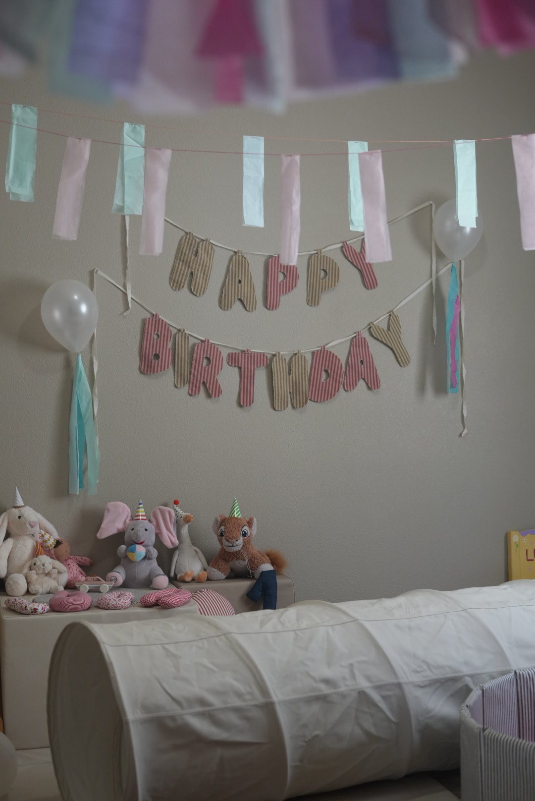 A cozy children's room scene decorated for a birthday, featuring a 'HAPPY BIRTHDAY' banner made of wooden letters with pink and beige striped letters, hanging above a bed with stuffed animals and balloons.