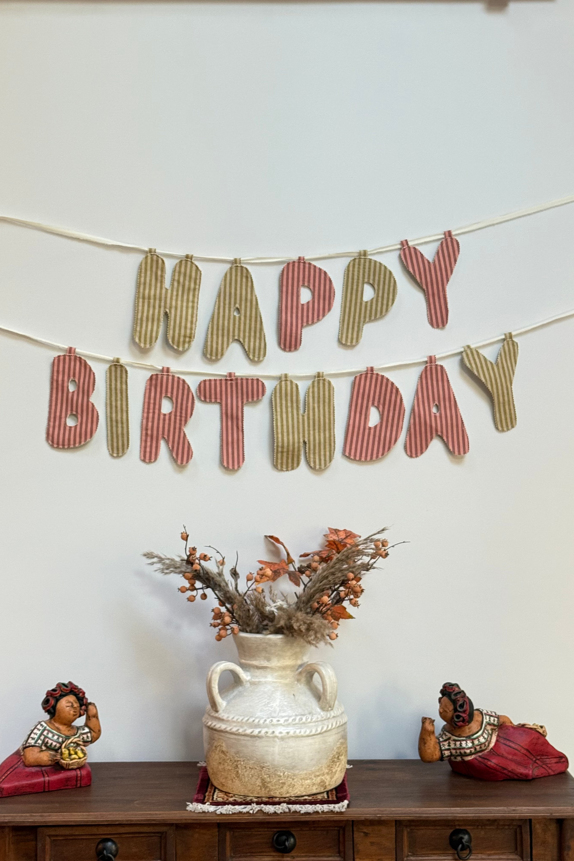 A rustic fabric 'HAPPY BIRTHDAY' banner hangs on a white wall above a wooden console table. The letters are made from striped green and muted red fabric, strung along thin cords.