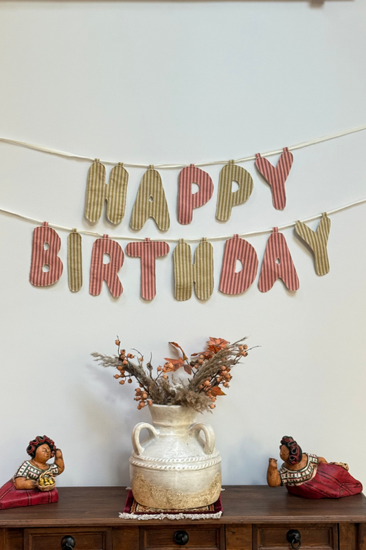 A rustic fabric 'HAPPY BIRTHDAY' banner hangs on a white wall above a wooden console table. The letters are made from striped green and muted red fabric, strung along thin cords.