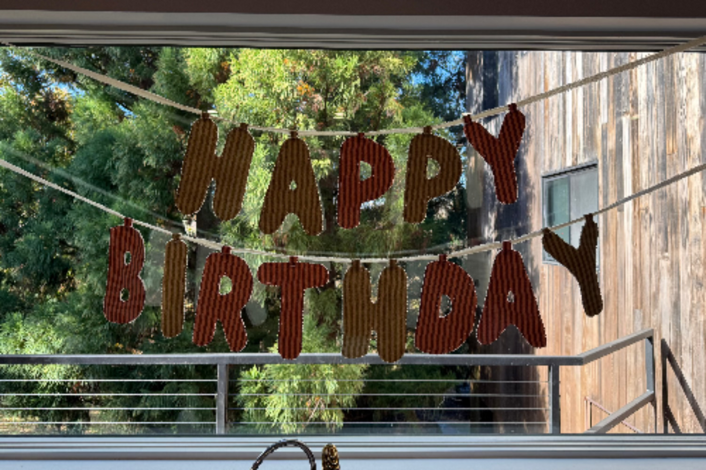 A 'Happy Birthday' letter banner made from reddish-brown wood-grain cutouts hanging on strings in front of a large window, with outdoor greenery and a wooden building visible in the background.